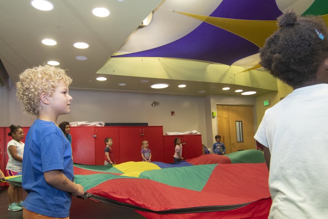 Young children standing in a circle and holding the edges of a parachute.
