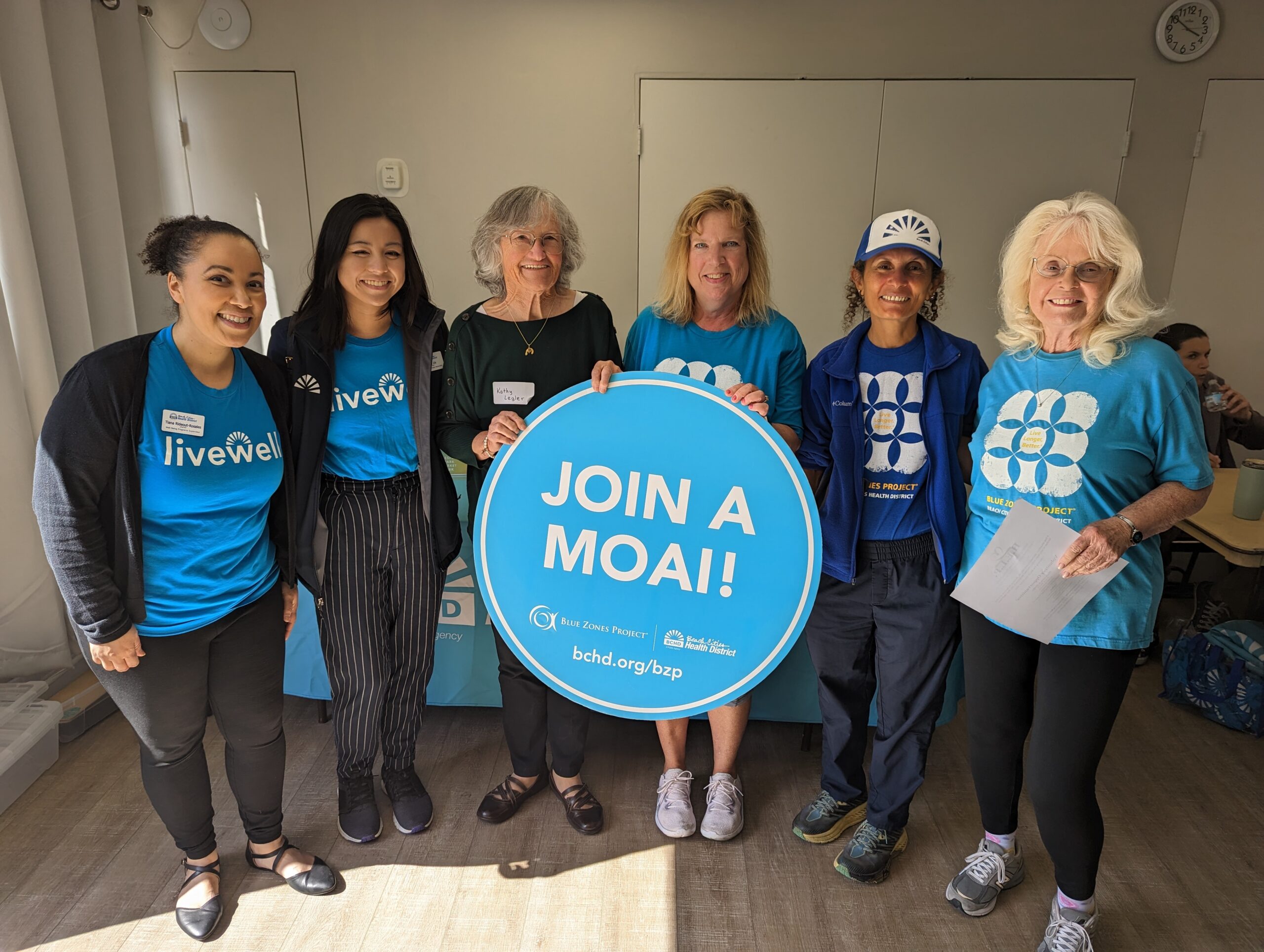 Six people stding in a row and the middle 2 persons are holding a round sign that reads "Join a Moai."