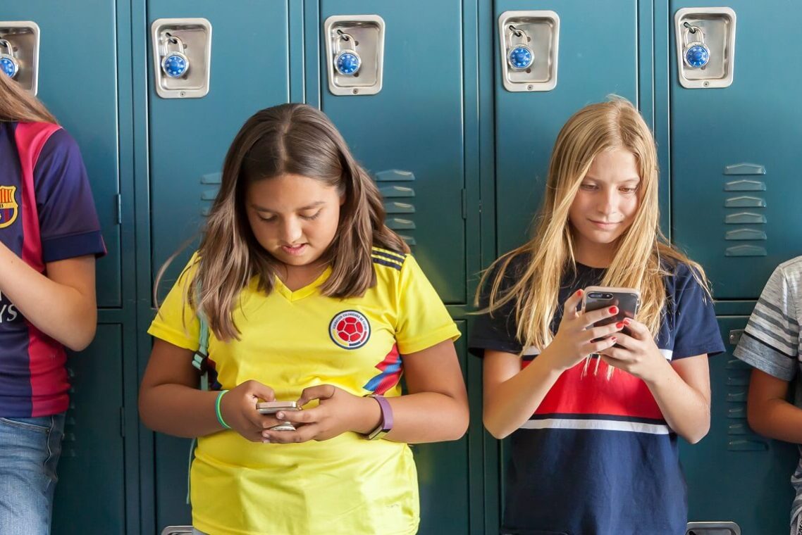 Four elementary aged students leaning against lockers, looking at their mobile devices.